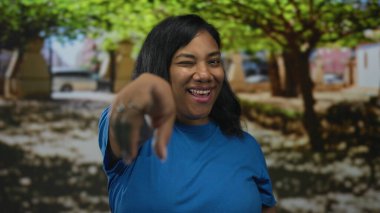 Mavi gömlekli kadın geniş bir gülümseme ve parktaki yeşil ağaçlar arasında parmak işareti; dostça bir davet.