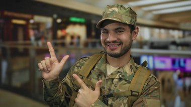 Kamuflaj üniformalı adam parmaklarını sağa sola gösteriyor ve alışveriş merkezi koridorunda gülümsüyor..