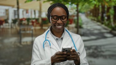 Kadın doktor dışarıda akıllı telefon kullanıyor. Restoran terasında dinleniyor. Güler yüzlü, açık hava ortamında teknoloji ve rahatlama karışımı bir şey sergiliyor..