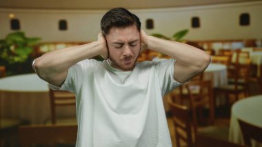 Sakallı genç adam gürültüyü engellemek için kalabalık bir restoranda kulaklarını kapatıyor, evde yuvarlak masalara ve tahta sandalyelere yaslanıyor, stresi ve rahatsızlığı vurguluyor..