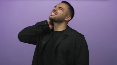 Siyah tişört giyen İspanyol genç adam mor stüdyoda boynuna dokunuyor gözleri kapalı ve gergin bir ifadeyle;.