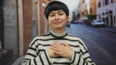 Şehir sokaklarında kısa saçlı bir kadın çizgili bir kazakla minnettar, elleri göğsüne bastırılmış, duygusal ve sakin bir şehir anı yaratıyor..