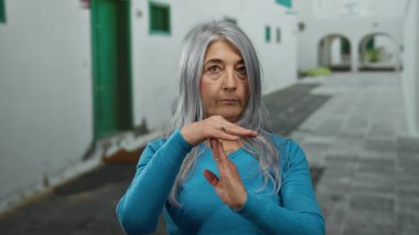 Gri saçlı yaşlı bir kadın, sessiz bir sokakta beyaz duvarlı ve yeşil kapılı geleneksel bir ortamda mola veriyor..