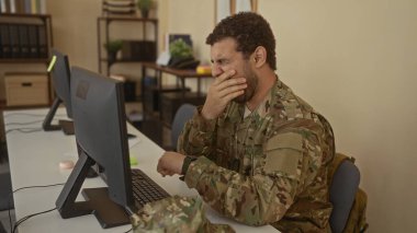 Odanın içindeki dağınık ofis masasında ağzını kapatan genç İspanyol asker monitörler ve dosyalarla dolu..