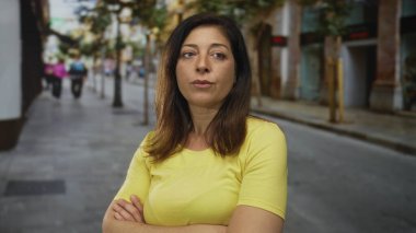 Kolları çapraz olan ve ağaçlarla ve mağazalarla dolu bir caddeye dik dik bakan bir kadın. Kararlılık..