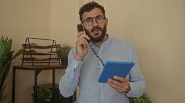 Ofiste telefon ve tablet kullanan İspanyol bir adam, arka planda bitki ve belgelerle birlikte çalışırken, iş ve iletişim temasını iletiyor.