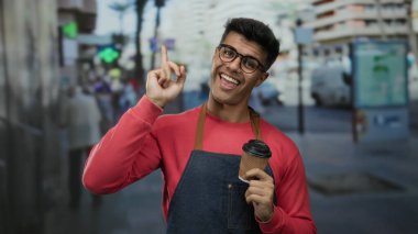 Gözlüklü ve önlüklü genç İspanyol bir adam şehrin işlek caddesinde kahve fincanı tutuyor. Dostane bir ifadeyle yukarı bakıyor. Şehir ortamında coşku ve açıklık olduğunu gösteriyor..