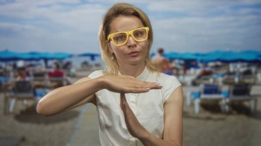 Sarı gözlüklü genç sarışın kadın bulanık şemsiyeler arasında kumlu plajda mola tabelası yapıyor..