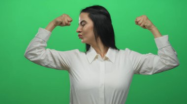 Beyaz tişörtlü İspanyol kadın, yeşil bir duvara yaslanmış kollarıyla kutlama yapıyor, yüz ifadeleri ve kendinden emin duruş ile neşe ve güç ifade ediyor..