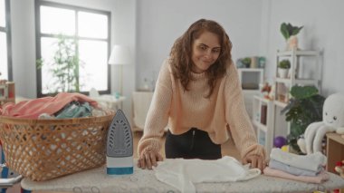 Çamaşır odasında gülümseyen bir kadın yakınlarda bir sepetle giysileri ütülerken modern ev işleri sergiliyor..