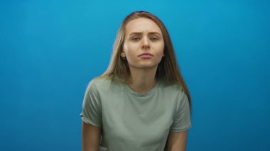 Canlı mavi bir arka plana karşı gelişigüzel giyinmiş bir kadın, izole edilmiş bir stüdyo ortamında merak ve içgözlem duygusu taşıyor..