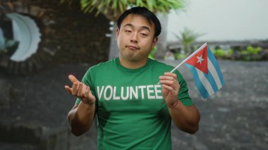 Şehir ortamında Küba bayrağı taşıyan genç bir adam, yeşil gönüllü tişört giyiyor, bir şehir ortamında uluslararası kültür veya gönüllü bağlantı öneriyor..