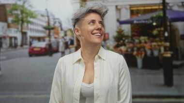 Kısa gri saçlı, gülümseyen yüzlü bir kadın işlek şehir caddesine bakıyor market tezgahlarıyla süslenmiş; iyimserlik.