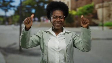 Gözlüklü bir kadın, yeşil bir caddede kendine güveni tam bir şekilde kendini gösteriyor. Açık renk bir ceket giyiyor ve hareketleriyle neşe saçıyor..