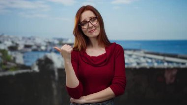 Deniz kenarındaki limanı işaret eden bir kadın, açık havada duruyor, kırmızı giyiyor, açık gökyüzü altındaki deniz ve teknelere karşı kendine güvenen bir ifade sergiliyor..