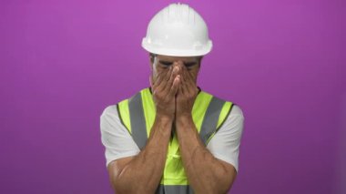 Beyaz kasklı ve yansıtıcı yelek giyen İspanyol adam mor stüdyoda elleri ile yüzünü örtüyor..