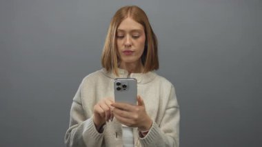 Gri arka planda akıllı telefon kullanan kadın, odaklı ifade ile günlük kazak giyiyor, bağlantıyı ve modern teknoloji eğilimlerini vurguluyor.