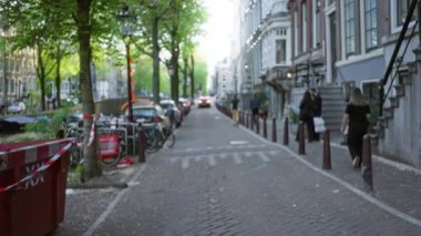 Dolaşan, araba kullanan, tarihi binaların ve yemyeşil ağaçların önünde sıralanmış bisikletlerin yer aldığı hareketli bir Amsterdam caddesinin bulanık görüntüsü.