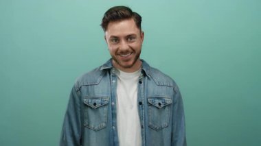 Sakallı, kot gömlekli genç adam, izole edilmiş yeşil arka plana gülümsüyor. Sıradan bir tarz ve pozitif enerji yansıtıyor.
