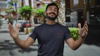 Sakallı İspanyol bir adam şehir caddesinde ağaçlar ve binalar arkasında mutluluk ve kendine güveni ifade ederek neşeli bir şekilde dolaşır..