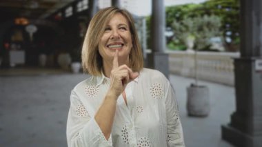 Beyaz bluzlu kadın sütunlarla kaplı bina koridorunda sessizlik için dudaklara parmak işareti yapıyor..