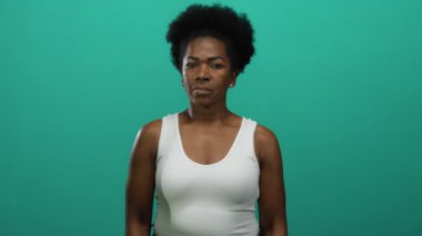 Yeşil arka planda başparmağıyla işaret eden bir kadın, izole bir ortamda jest yapıyor, genç kadının odaklanmış ifadesini yakalıyor, beyaz kolsuz bluz giyiyor..