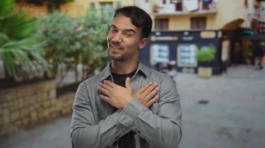Genç bir adam, İspanyol asıllı, açık havada gülümsüyor, kolları çaprazlanmış bir şekilde şehir merkezinde binaların arka planında, mutluluğu ve rahatlığı aktarıyor..