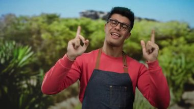 Önlüklü ve gözlüklü genç adam gülümsüyor ve canlı yeşil bir açık hava parkında dikiliyor..