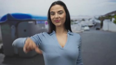 Sokakta gülümseyen ve poz veren bir kadın parmaklarıyla barış işareti yapıyor, mavi bir gömlek giyiyor, sıradan bir şehir ortamında mutluluk ve neşe taşıyor..