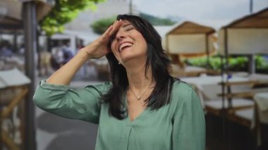 Yeşil bluzlu orta yaşlı bir kadın, parlak güneş ışığı altında terasta elini açık tutarak alnına dokunur..