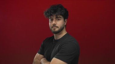 Sakallı İspanyol genç adam kollarını parlak kırmızı arka plana yaslıyor. Ciddi görünüyor ve gülümsüyor..
