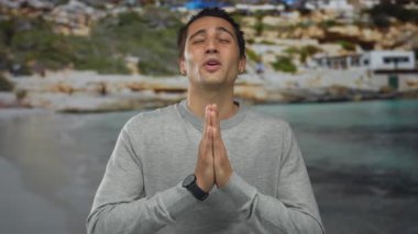 Genç adam deniz kenarında sakin bir ifadeyle dua ediyor, gözleri kapalı, gri kazak giyiyor, yakın plan çekimi, huzurlu plaj arka planı, yaz havasını, açık havayı ima ediyor..