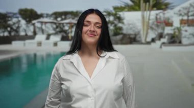 Havuz başında sessiz hareketler yapan bir kadın, berrak bir gökyüzünün altında, beyaz dekor, canlı yeşillik ve mavi suyla huzurlu, lüks bir tatil ortamı sergiliyor..