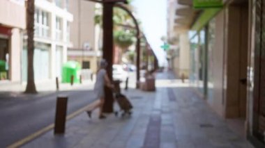 Açık bir sokakta, güneşli bir günün altında palmiye ağaçları ve dükkanlarla çevrili bir arabanın altında yürüyen bir kadının bulanık görüntüsü.