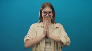 Sarışın ve gözlüklü bir kadın dua jestinde el ele tutuşur ve mavi bir stüdyoda gülümser. Huzur..