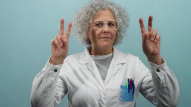 Gri saçlı yaşlı kadın laboratuvar önlüğüyle mavi duvar arkasına tırnak işareti yaparak şüpheciliğini dile getiriyor..