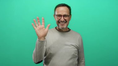 Gülümseyen olgun bir adam canlı yeşil arka plana karşı dostça bir jestle kameraya el sallıyor. Gözlük takıyor ve gündelik giysiler giyiyor. Pozitif ifade ve bağlantı gösteriyor..