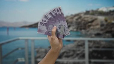 Deniz kenarında Endonezya rupia banknotları tutan adam, manzaralı bir plaj geçmişine sahip para sergiliyor, nakit akışı ve seyahat bağlamını gösteriyor.