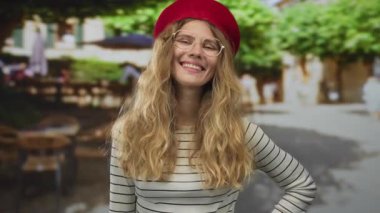 Kadın portresi, bir sokak kafesi sahnesinde gülümseyen parlak bir yüz yakalıyor. Sarışın bir ev sahibesi, açık hava ortamının tadını çıkarıyor. Gözlüklü ve bereli, yemyeşil çerçeveli..