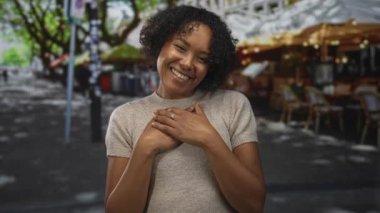 Açık kafe masalarının yakınındaki güneşli şehir caddesinde, yeşil ağaç tepe örtüsünün altında el ele tutuşarak gülümseyen kadın; minnettarlık.