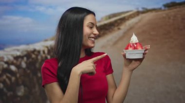 Genç İspanyol kadın sevinçle üzerinde çilek olan lezzetli dondurmayı işaret ediyor mavi gökyüzünün altında manzaralı bir zemin üzerinde, heyecan ve neşe ifade ediyor..