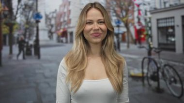 Beyaz noktalı uzun saçlı sarışın kadın şehir caddesinde kameraya parmak hareketi yapıyor..