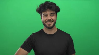 Sakallı gülümseyen genç bir adam neşeli ve kendinden emin bir ifadeyle olumlu ve olumlu bir görünüm sergileyen yeşil bir geçmişe başparmak sallıyor..