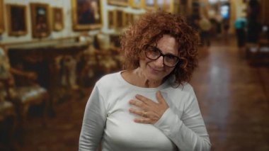 Orta yaşlı esmer bir kadın sanat galerisinde gülümsüyor, gözlük takıyor ve beyaz bir gömlek giyiyor..