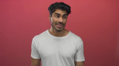 Beyaz tişörtlü bir adam kırmızı stüdyoda başını işaret ediyor. Tereddütlü bir ifadeyle, düşünceli bir ruh hali içinde..