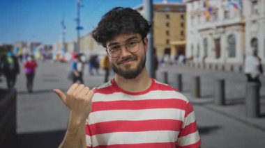 Sakallı, gözlüklü ve çizgili tişörtlü genç İspanyol adam güneşli bir günde açık havada başparmak uçlarını gösteriyor..
