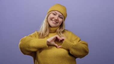 Sarı süveter ve mor arka planda bereyle gülümseyen, sevgi ve pozitifliği ifade eden kalp hareketi yapan bir kadın..