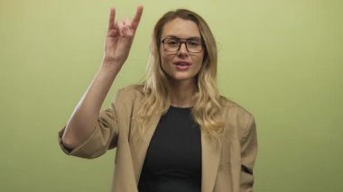 Yeşil arka planda gözlüklü ve gülümseyen bir kadın el hareketi yapıyor..