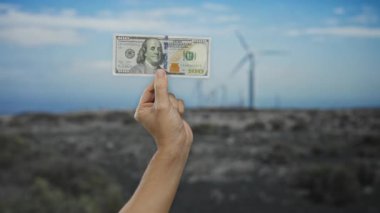 Adam arka planda yel değirmenleri olan bir alanda Amerikan doları tutuyor, finans ve sürdürülebilir enerji arasındaki ilişkiyi sembolize ediyor..