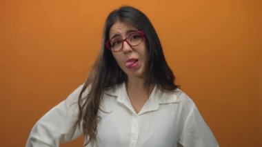 Gözlüklü bir kadın turuncu arka planda neşeli yüzler sergiliyor beyaz gömlek giyiyor ve izole ortamda mizah ve ifade gösteriyor..
